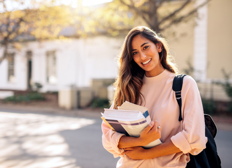 A smiling young woman holding several books, radiating joy and enthusiasm.